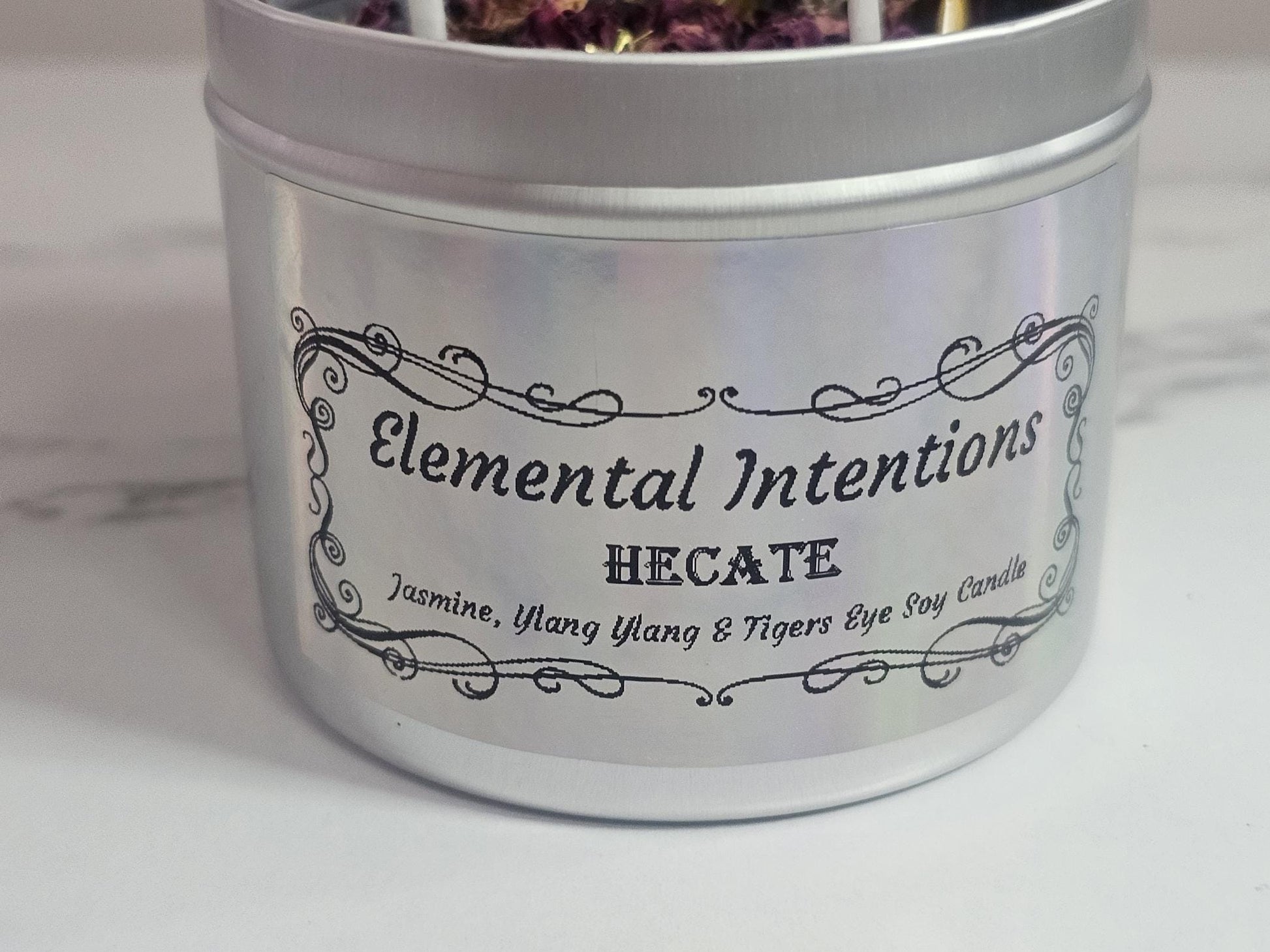 A front shot of a container candle in silver with a holographic label which says &#39;Elemental Intentions&#39; and &#39;Hecate&#39;. It is labelled as a Jasmine, ylang ylang and tigers eye soy wax candle.