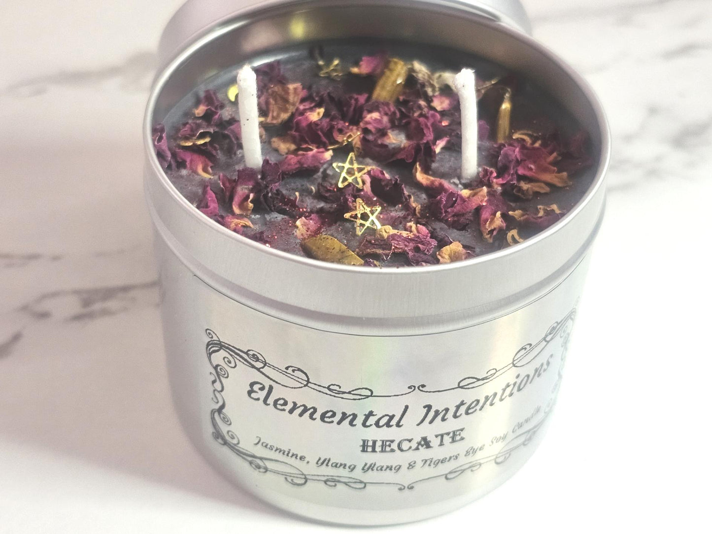 A silver container candle with a luminescent silver label saying &#39;Elemental Intentions, and Hecate&#39;. Label states the scent is Jasmine, Ylang Ylang and it has tigers eye crystals as decorative toppers with rose petals and gold moon and stars decorative pieces along with two white cotton wicks. There is a silver lid to the back which goes on the top and it&#39;s on a white marble background.