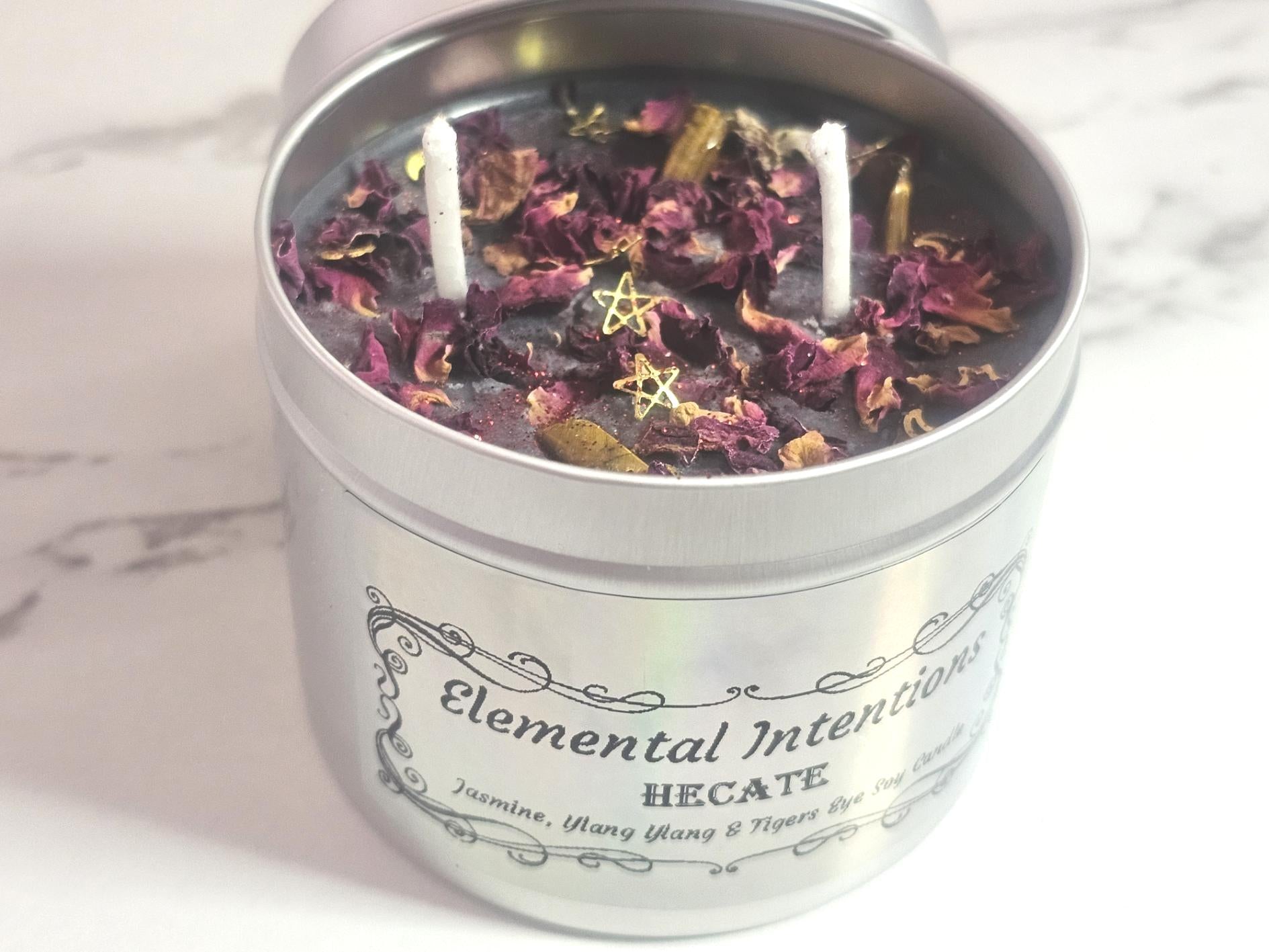 A silver container candle with a luminescent silver label saying &#39;Elemental Intentions, and Hecate&#39;. Label states the scent is Jasmine, Ylang Ylang and it has tigers eye crystals as decorative toppers with rose petals and gold moon and stars decorative pieces along with two white cotton wicks. There is a silver lid to the back which goes on the top and it&#39;s on a white marble background.