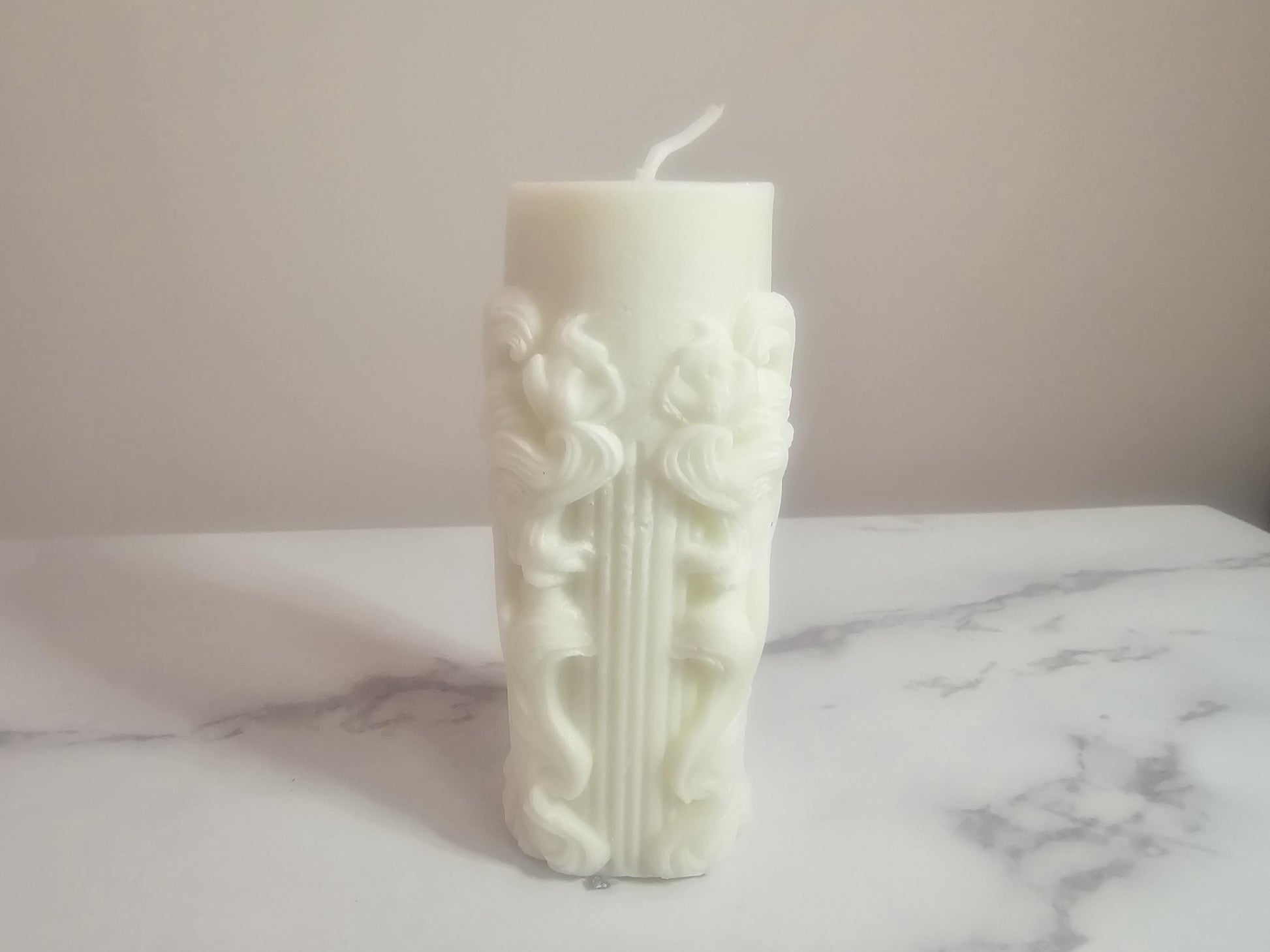 A white pillar candle with a white cotton wick and a floral framed column design etched all the way around.