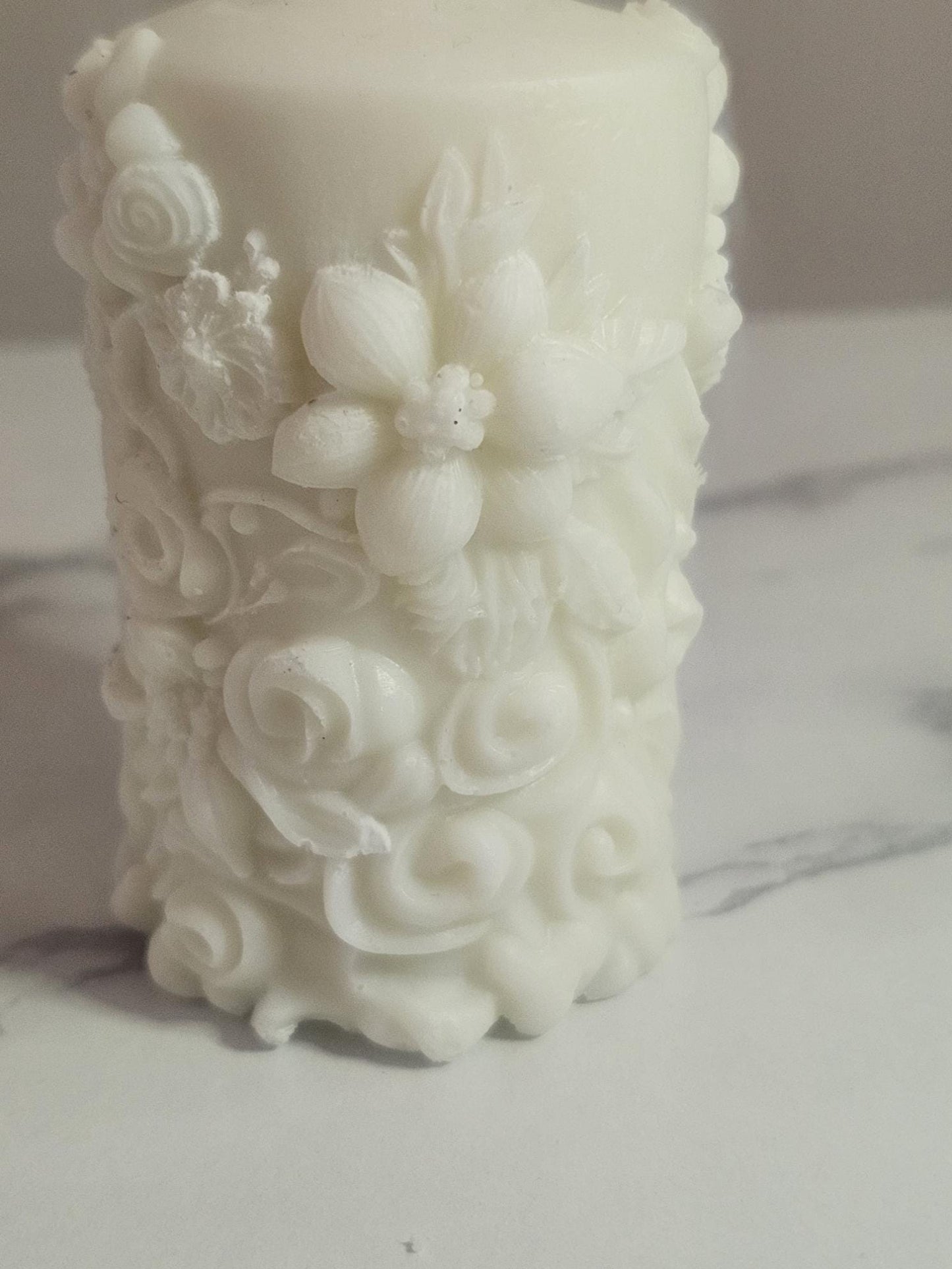 A white soy wax pillar candle adorned in floral 3D etchings all the way around with flowers, roses and leaf design and a cotton wick.