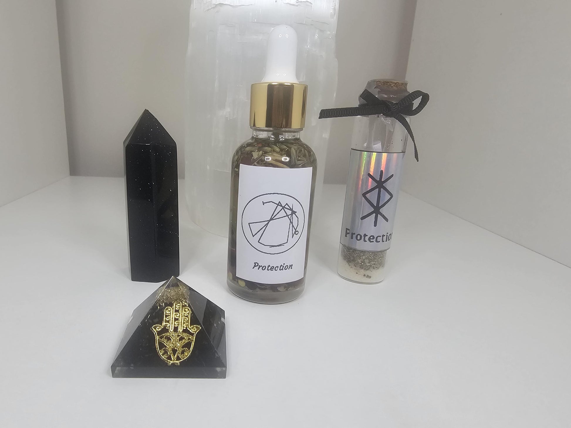 A small clear dropper bottle with a gold lid, filled with protective herbs and with a sigil standing for protection on the front of the bottle. The bottle stands next to a black and gold hamza pyramid, a black obsidian crystal tower and a small glass protection spell jar.