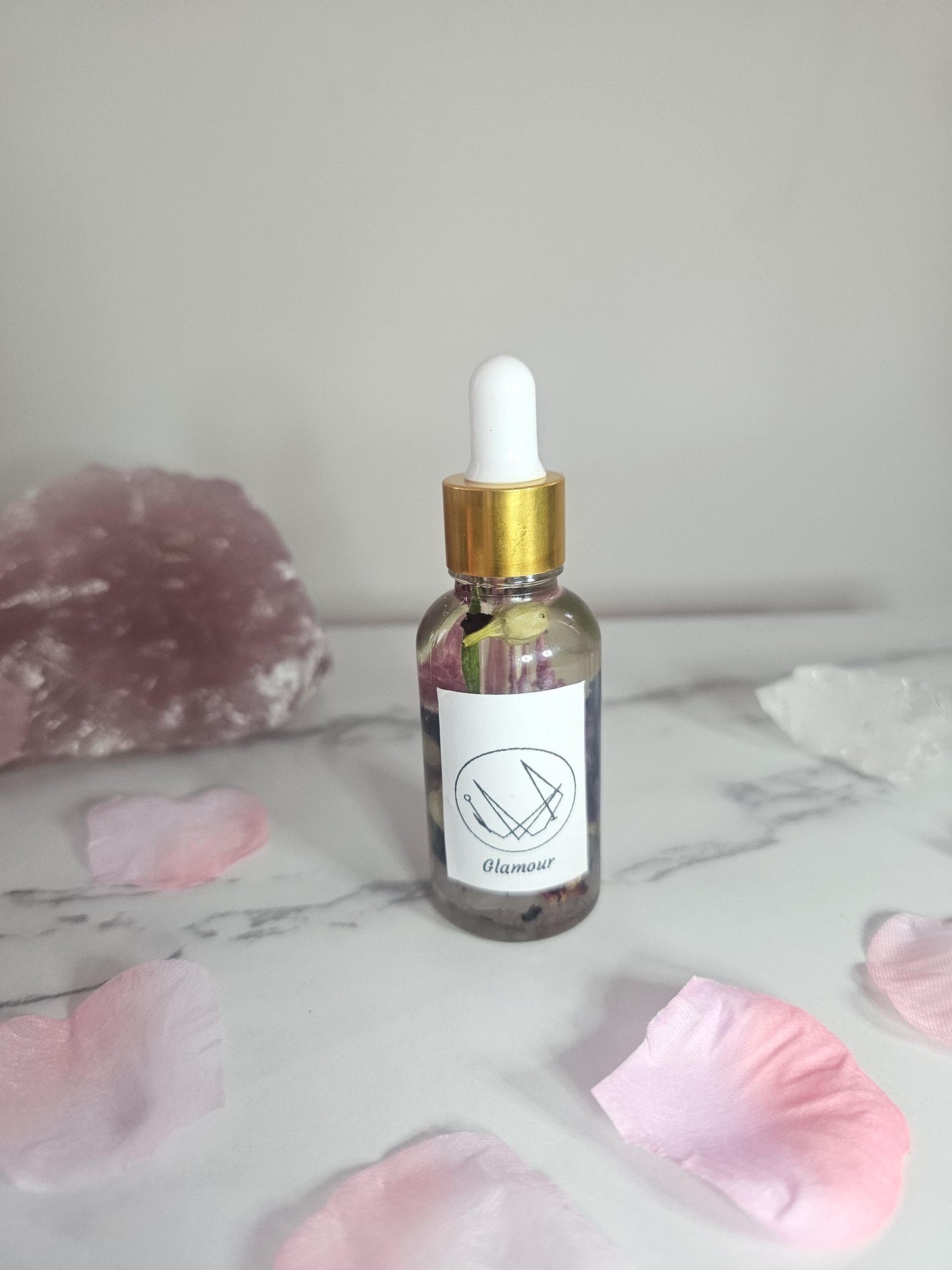 A clear dropper bottle with a gold top filled with oil and pink flowers and herbs. There is a label on the front with a sigil and the word &#39;glamour&#39; and it is surrounded by pink petals and a rose quartz stone.