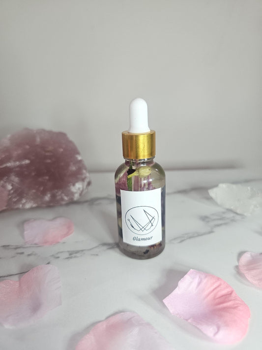 A clear dropper bottle with a gold top filled with oil and pink flowers and herbs. There is a label on the front with a sigil and the word &#39;glamour&#39; and it is surrounded by pink petals and a rose quartz stone.