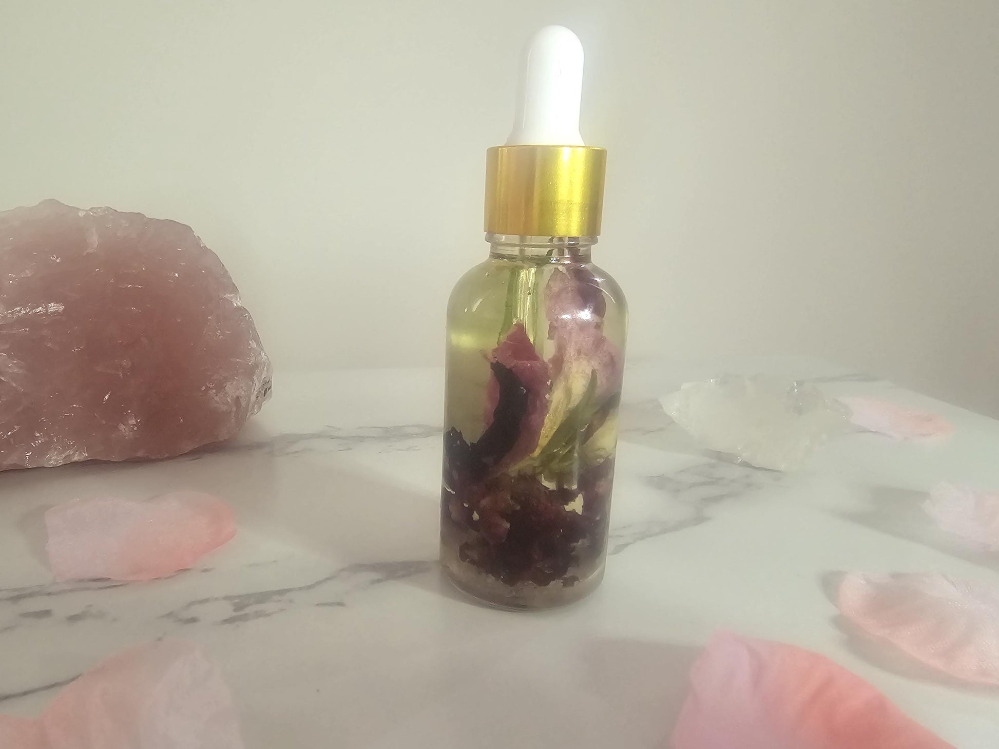 A clear dropper bottle with a gold top, filled with glamour oil. There are pink petals surrounding it and a rose quartz crystal.