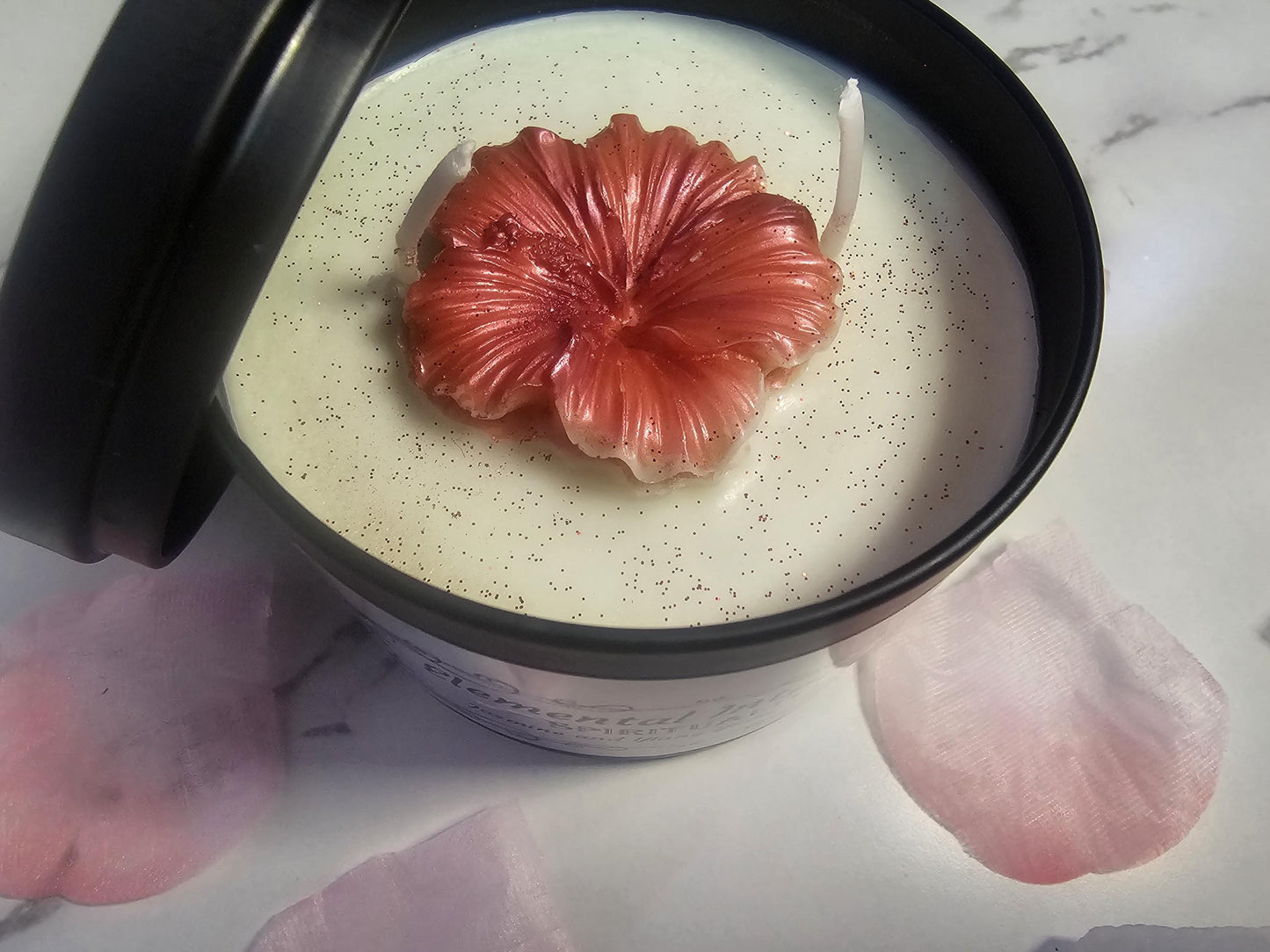 A black candle container on a white background, filled with white soy wax and topped with a coral pink wax peony. There are shimmers of coral glitter on top and it is surrounded by pink flower petals.