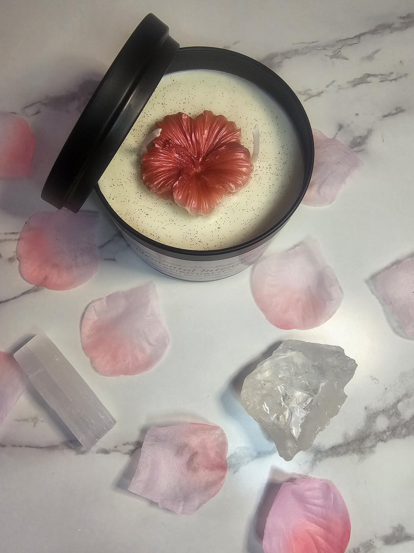 An above shot of a black candle tin surrounded by pink flower petals and clear crystals. The tin is filled with white soy wax and it is topped with a coral pink wax peony flower.