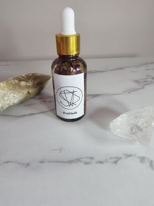 A clear dropper bottle with a gold top and a label on the front with a sigil, that also says &#39;gratitude&#39;. The bottle is filled with clear oil, flowers and herbs and is surrounded by crystals.