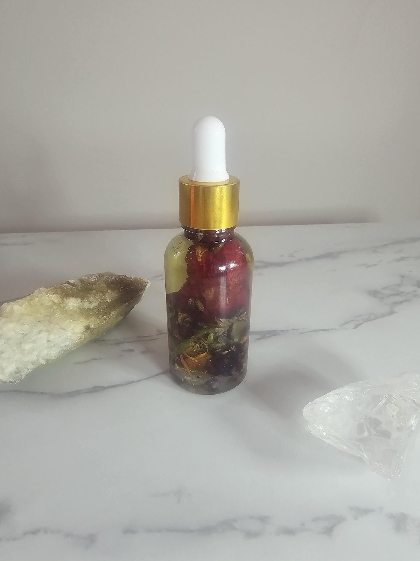 A clear glass dropper bottle with a gold top on a white background, surrounded by natural crystals. The bottle is filled with clear oil and red and orange flower petals, as well as herbs and small crystals.