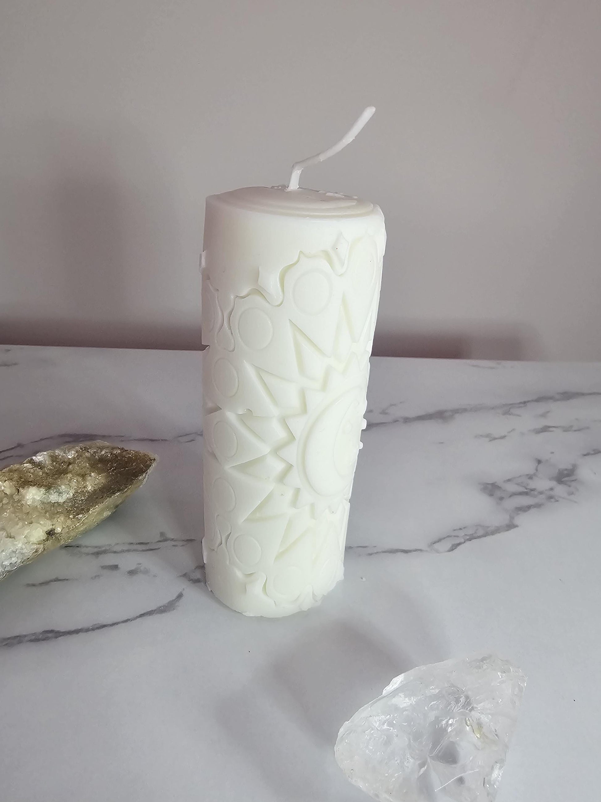 A tall white altar candle with an etched stars and moon design on a white marble background and surrounded by crystals.