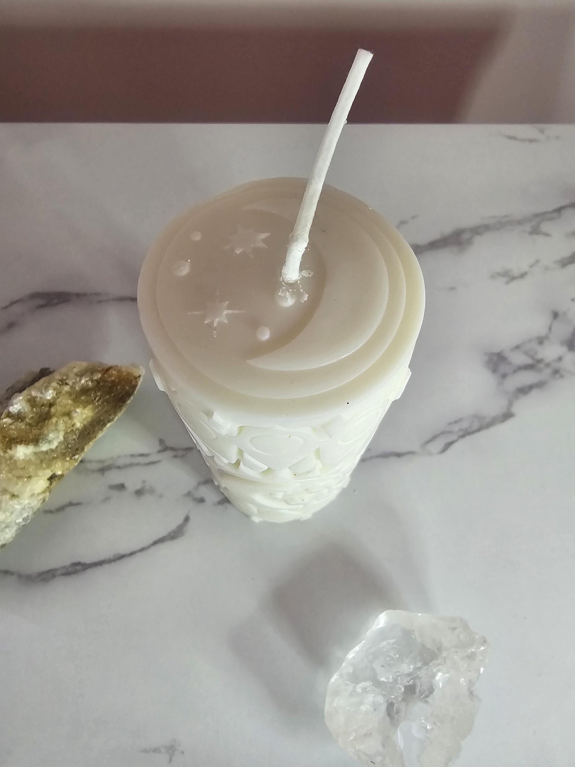 An above shot of a tall white pillar candle etched with a 3D design of moon and stars with a white wick on a marble background.
