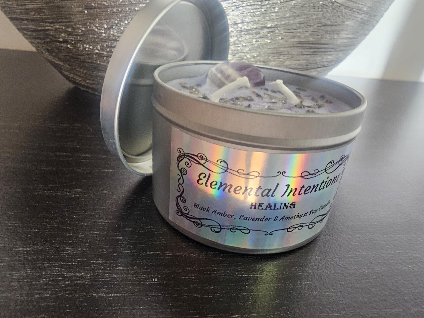 A silver candle container with a silver lid behind it and a ornamental holographic ticker framed in black saying &#39;elemental intentions&#39;, &#39;healing&#39; and labelled as a Black Amber, Lavender and Amethyst Soy Candle.