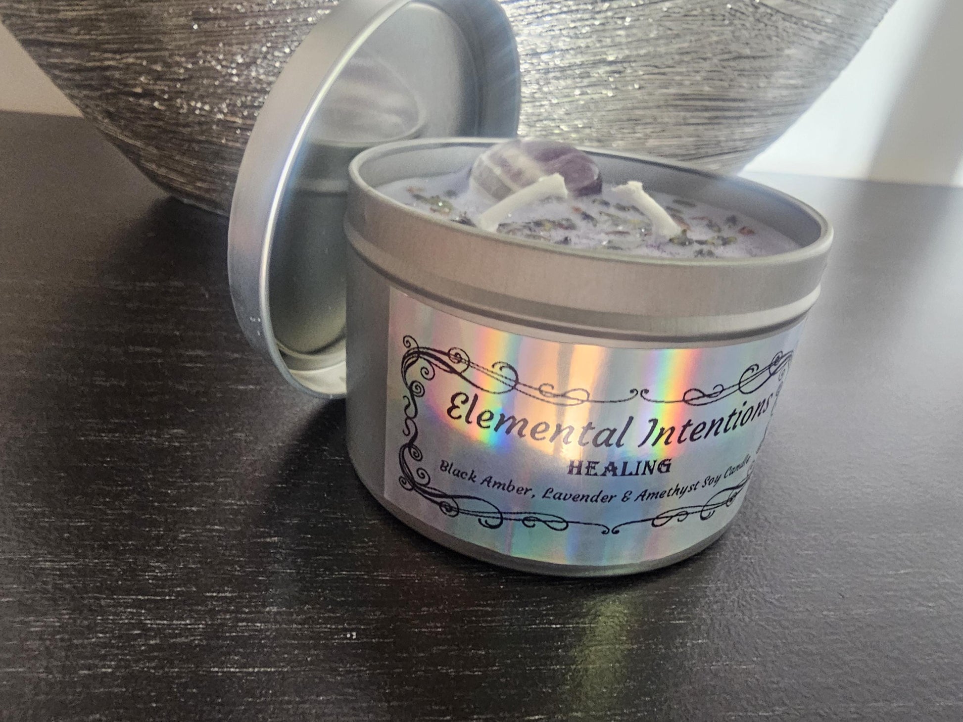 A silver candle container with a silver lid behind it and a ornamental holographic ticker framed in black saying &#39;elemental intentions&#39;, &#39;healing&#39; and labelled as a Black Amber, Lavender and Amethyst Soy Candle.