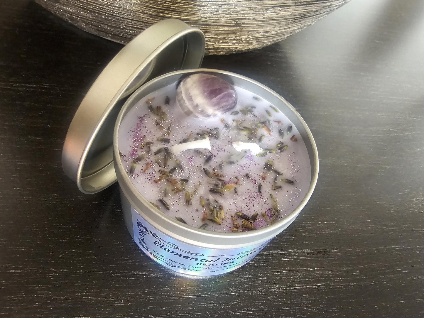 An above shot of a silver container candle filled with light purple wax and topped with lavender, purple amethyst and purple glitter, on a black background.