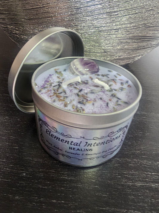 A silver candle container with a li beside it filled with light purple soy wax and decorated with purple amethyst and lavender, and shimmering with purple candle glitter. Has two white cotton wicks on a black background.