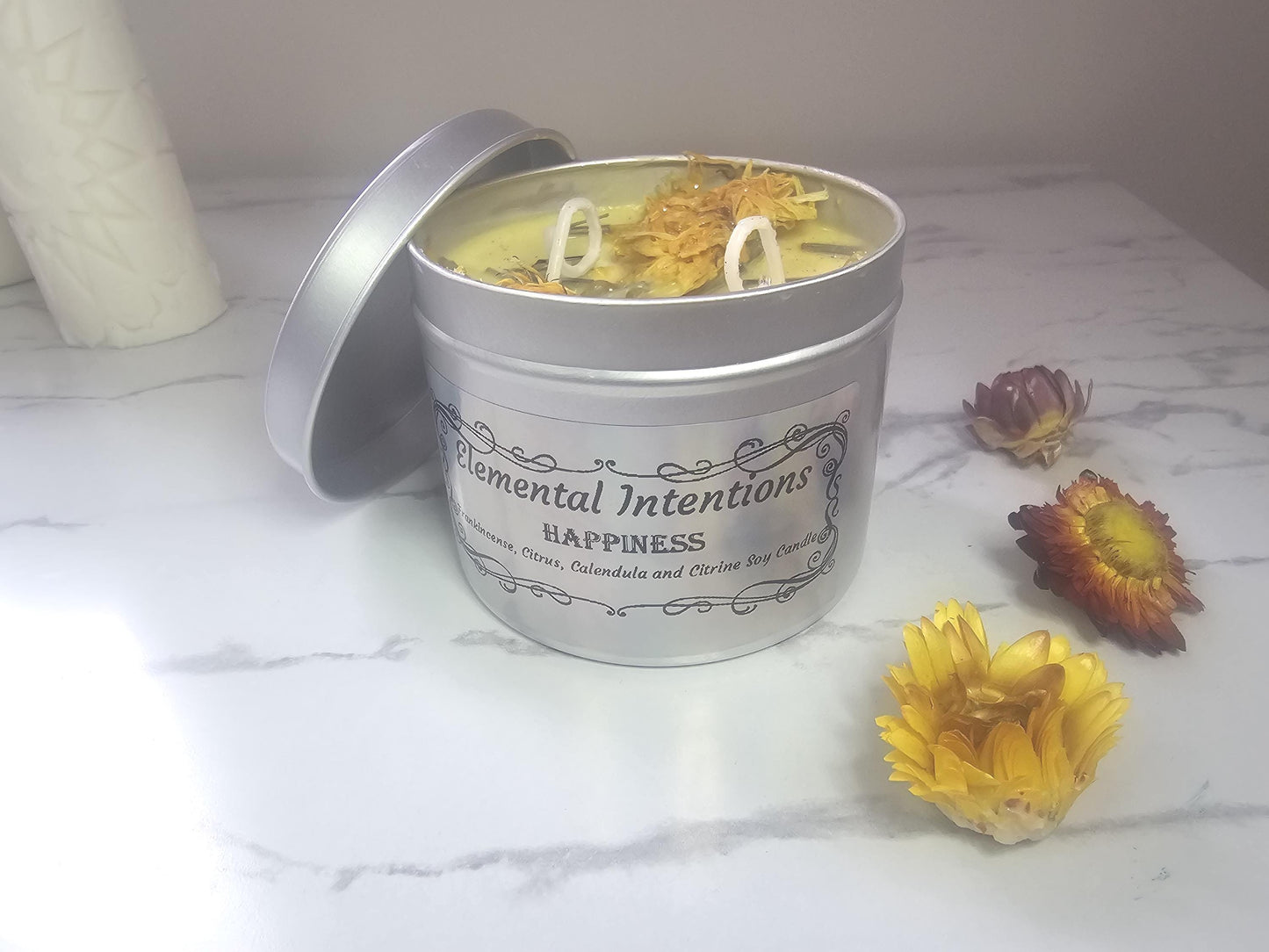 A silver container candle filled with yellow soy candle wax and topped with calendula flowers and citrine crystals. The holographic label on the front reads &#39;elemental intentions&#39;, &#39;happiness&#39; and &#39;frankincense, citrus, calendula and citrine soy candle&#39;.