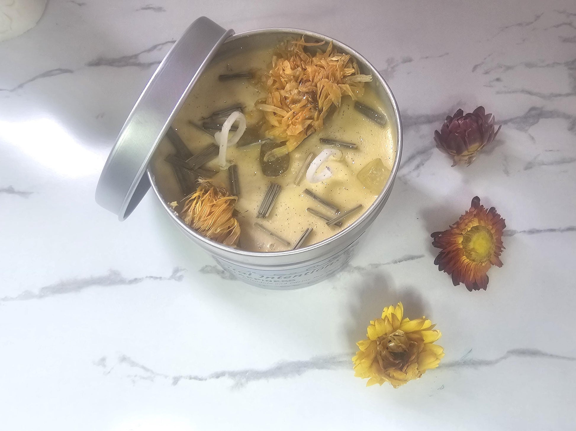 An above shot of a yellow candle in a silver container topped with decorative calendula, lemongrass and citrine crystals, which is surrounded by yellow and red flowers on a white marble background.