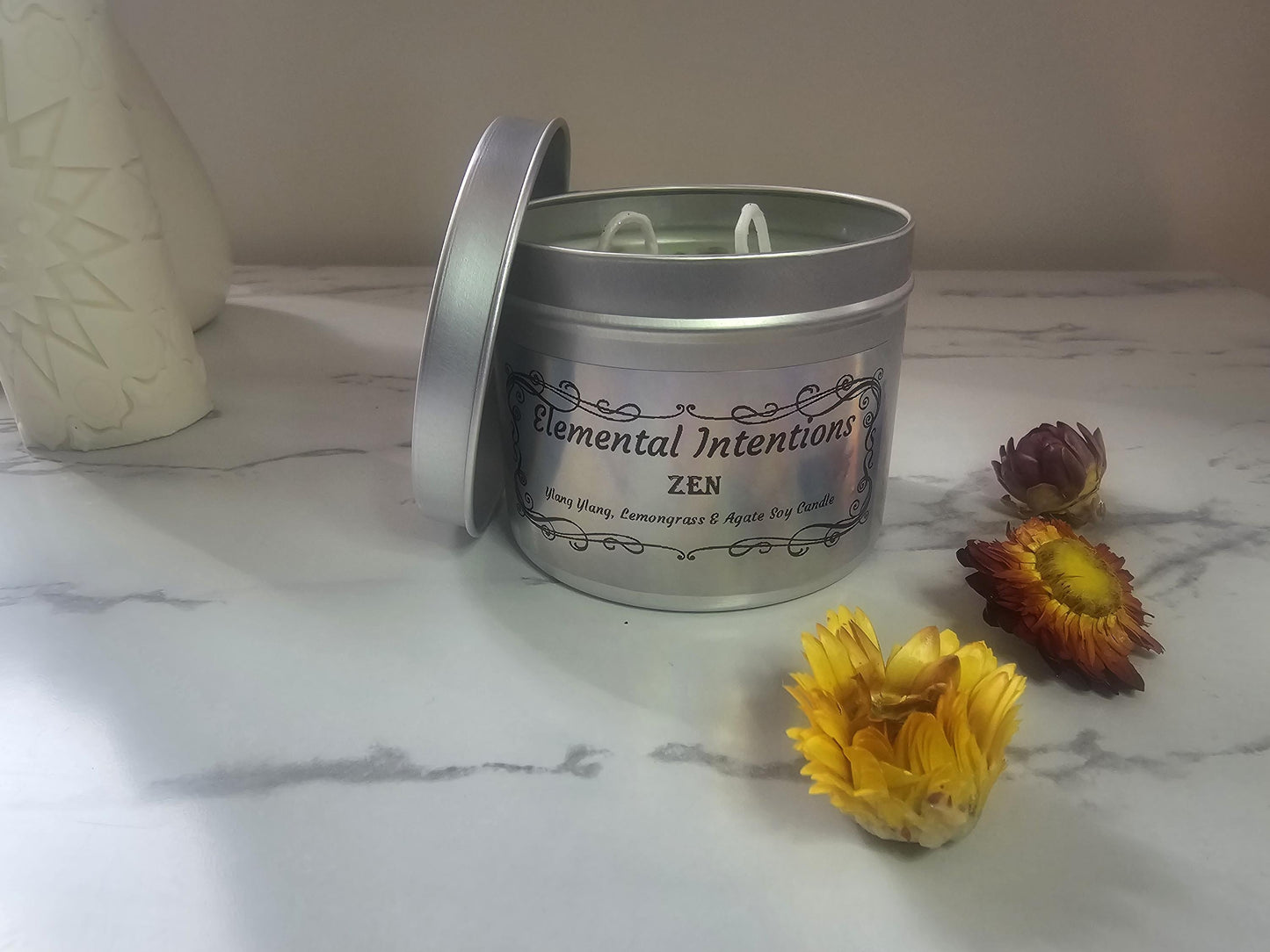 A front shot of  silver container candle with a holographic label that says &#39;elemental intentions&#39;, &#39;zen&#39; and &#39;ylang ylang, lemongrass and agate soy candle&#39;.