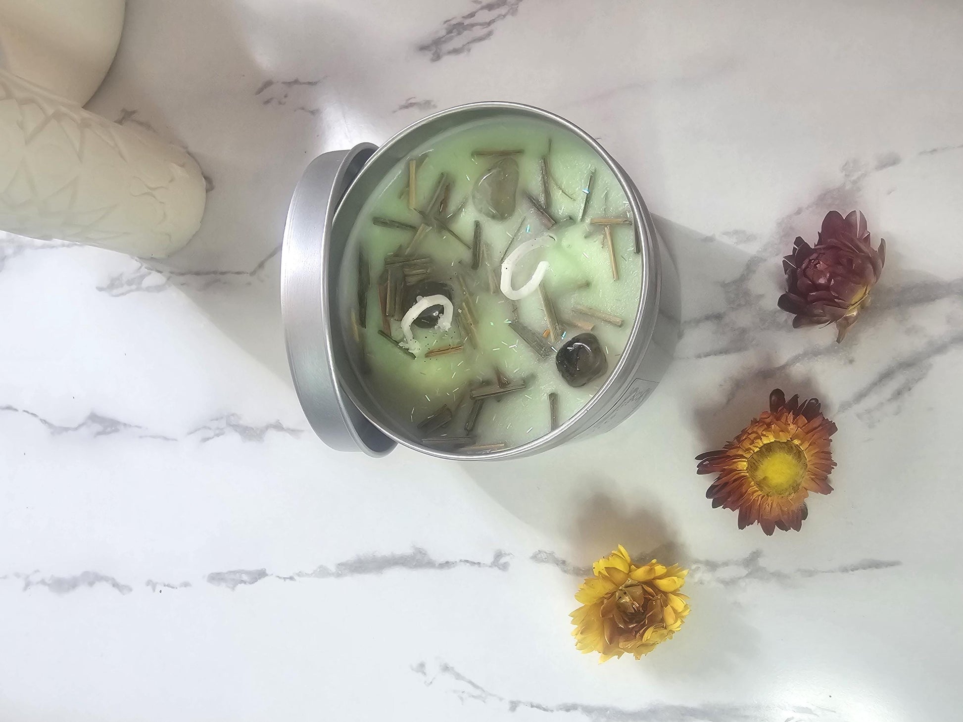 An above shot of a light green candle in a silver container surrounded by chrysanthemums. The candles is topped with pieces of decorative lemongrass, small pieces of glitter and small pieces of agate crystals.