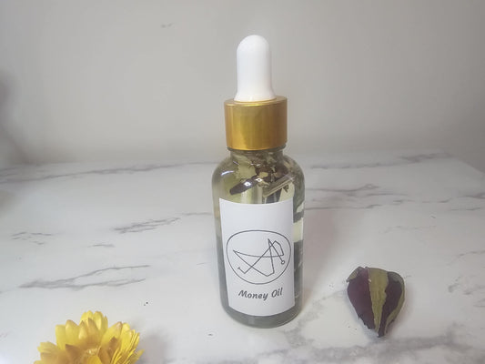 A clear dropper bottle with a gold top on a marble white background surrounded by red and yellow flowers. The front white label says &#39;money oil&#39; and it is filled with clear oil and herbs.