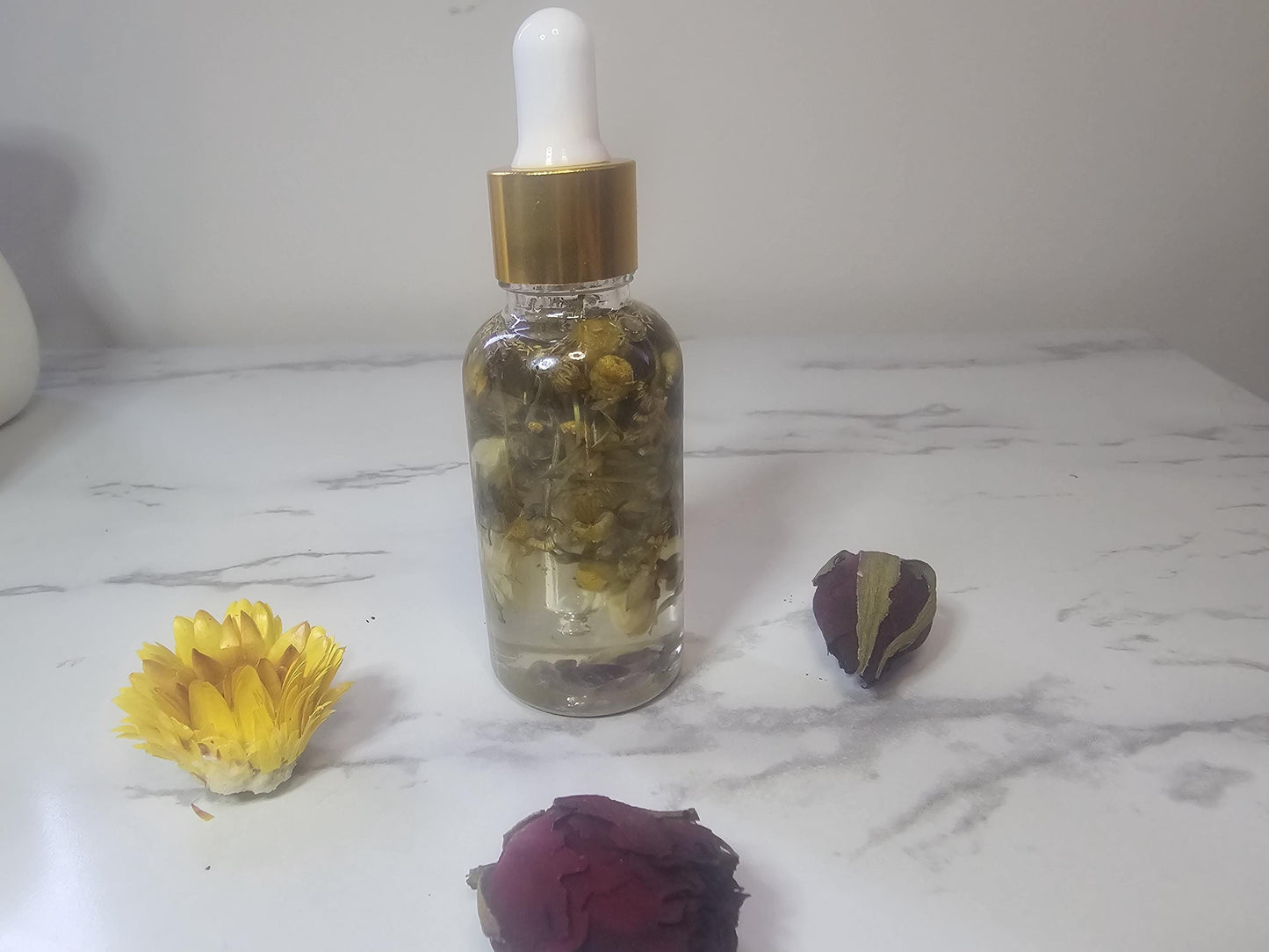 A clear glass dropper bottle with a gold top on a white marble background. the bottle is filled with clear oil and contains different varieties of dried herbs.