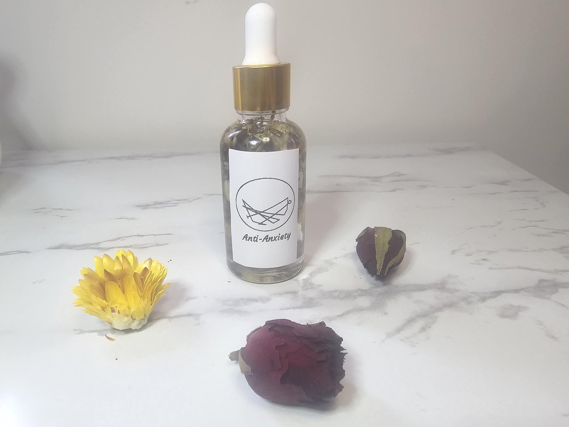 A clear glass dropper bottle with a gold top on a white marble background surrounded by red and yellow dried flowers. The front label is white and contains a picture sigil and the words &#39;Ant-anxiety Oil&#39;.