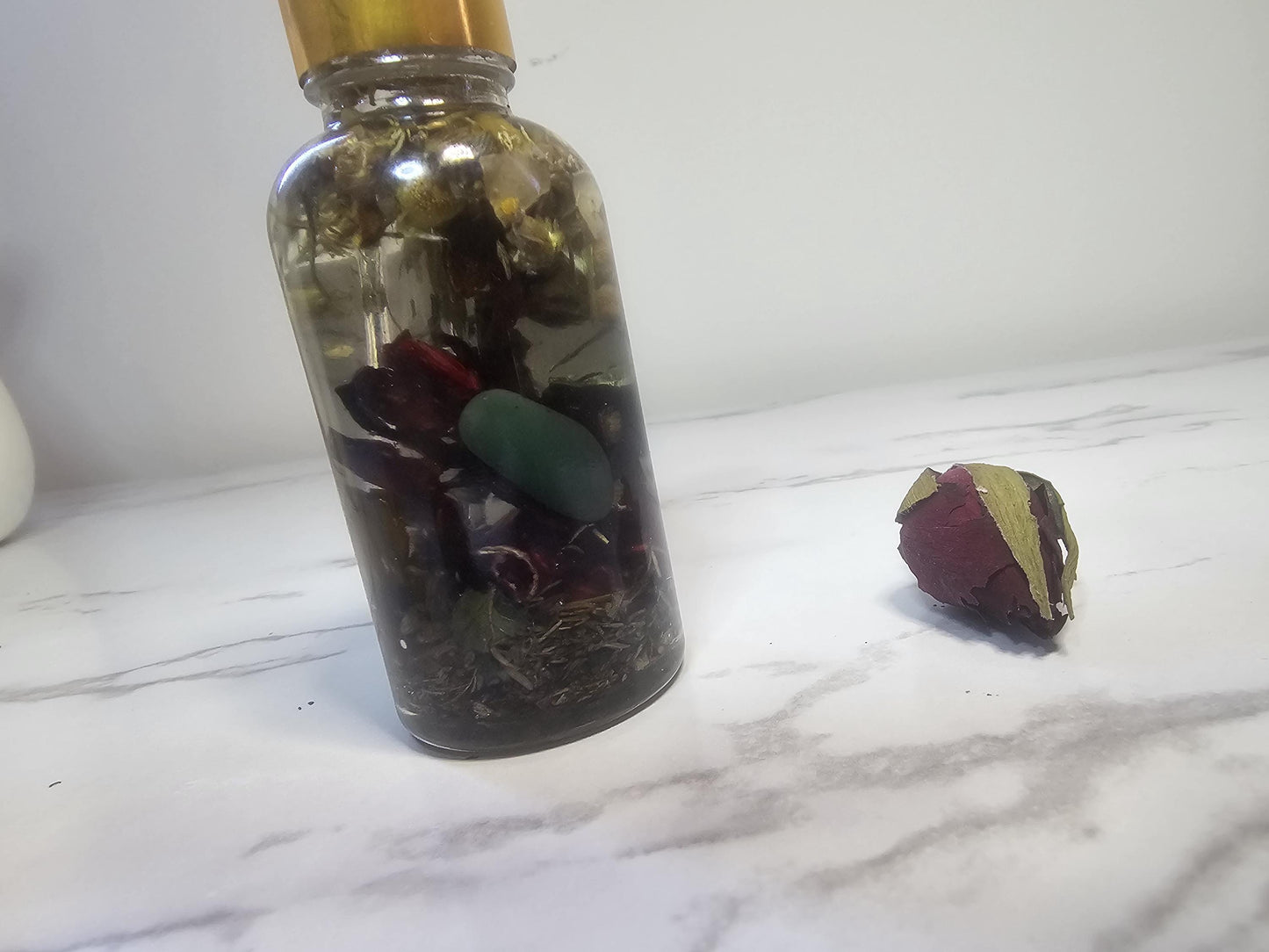 A clear glass dropper bottle with a gold top on a white marble background surrounded by red and white dried flowers. The bottles is filled with clear oil, herbs, and various coloured crystals such as aventurine and tigers eye.