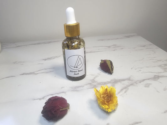 A clear glass dropper bottle with a gold top on a white marble background surrounded by red and white flowers. There is a white label on the front with a picture sigil and the words &#39;attract good luck oil&#39;.