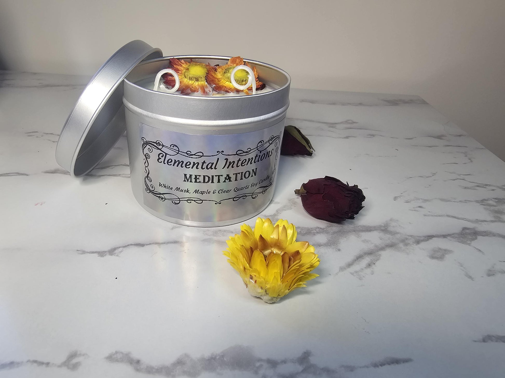A silver candle tin with a holographic label that says &#39;elemental intentions&#39;, &#39;meditation&#39; and &#39;white musk, maple and clear quartz soy candle&#39;. There are red and yellow chrysanthemums topping the light green wax.