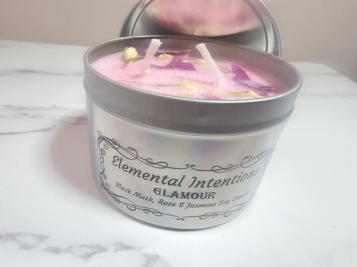 A silver candle container with a silver holographic label containing an ornate black frame and the words &#39;elemental intentions&#39; and &#39;glamour on the front. The candle is made of pink soy wax and two cotton wicks and decorated with lotus petals and jasmine buds.