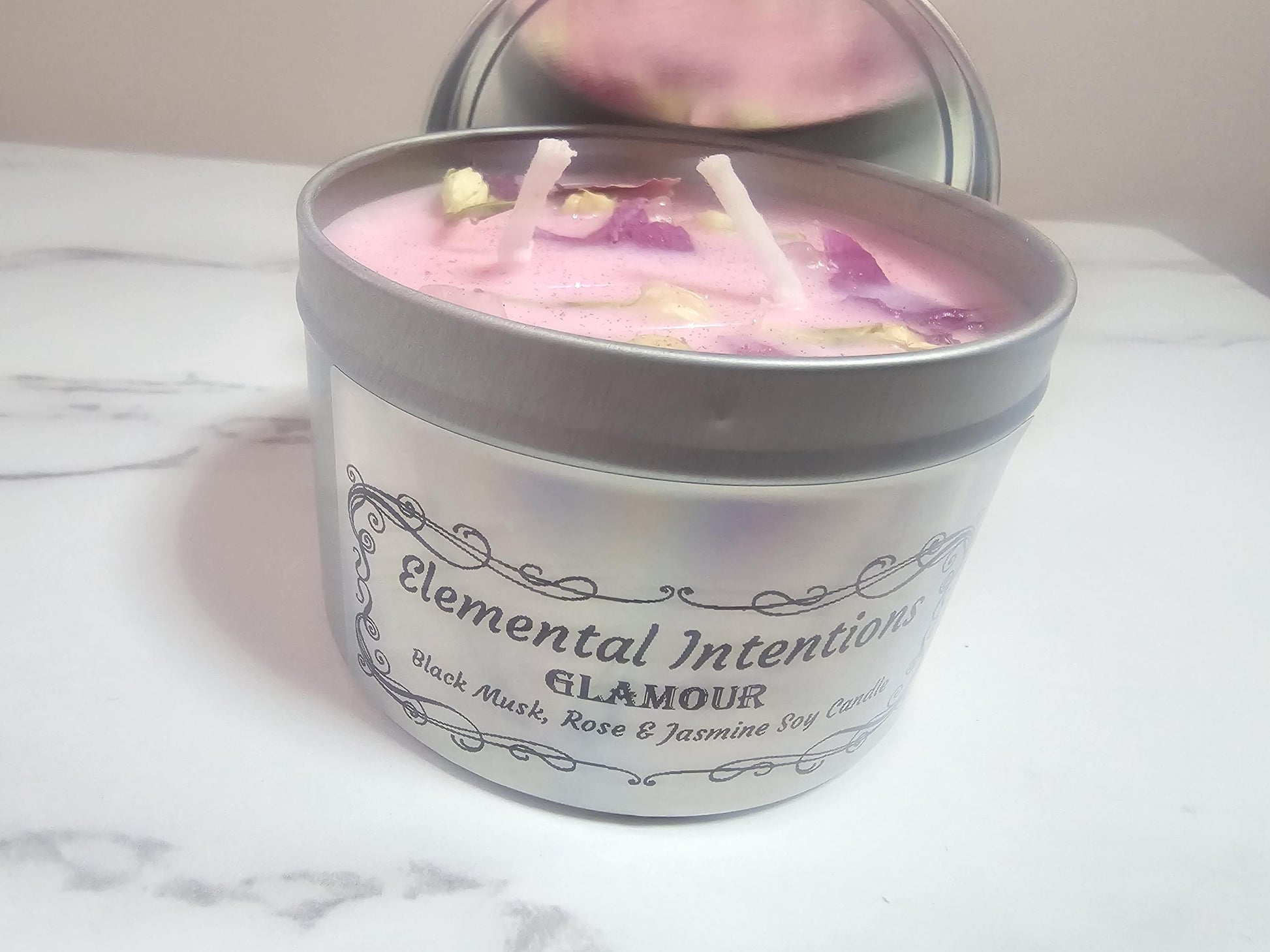 A silver candle container with a silver holographic label containing an ornate black frame and the words &#39;elemental intentions&#39; and &#39;glamour on the front. The candle is made of pink soy wax and two cotton wicks and decorated with lotus petals and jasmine buds.