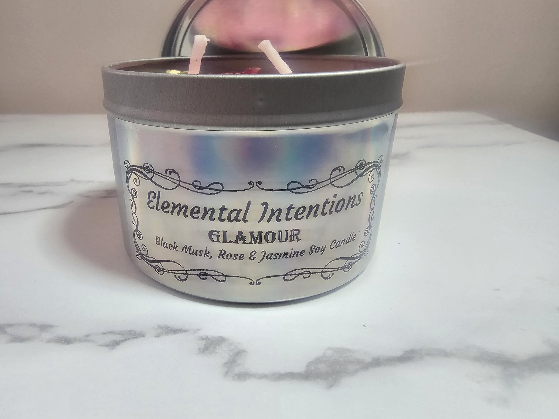 A frontal shot of a silver candle container with a holographic label with black ornate framing. The label reads &#39;elemental intentions&#39;, &#39;glamour&#39; and the words &#39;black musk, rose and jasmine soy candle&#39;.