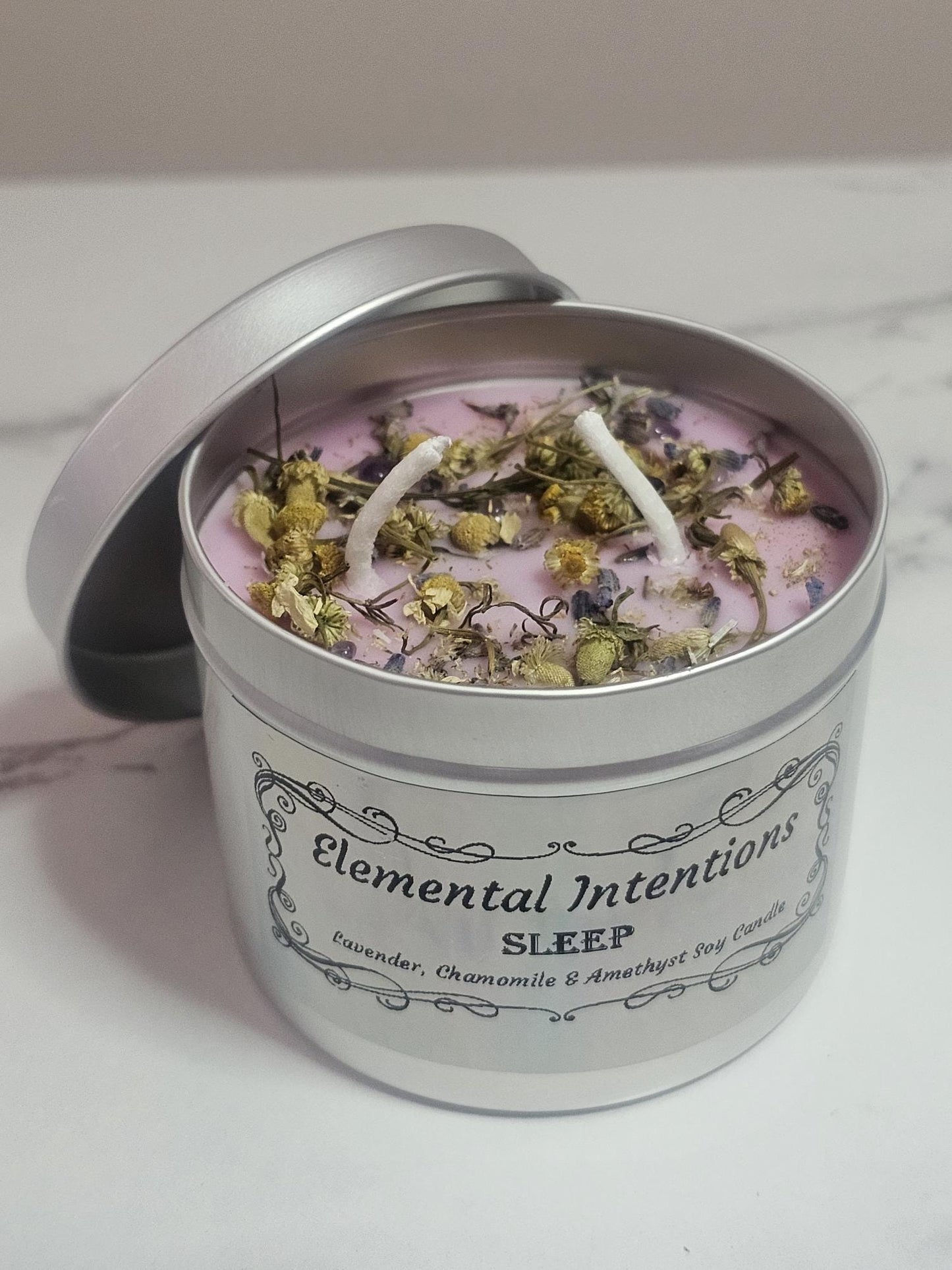 A silver candle container on a white marble background with a silver lid behind it. Inside is a light purple candle with two cotton wicks topped with decorative chamomile pieces and small amethyst crystal chips. The label says &#39;Elemental Intentions&#39; &#39;Sleep&#39; and it is labelled as a Lavender, Chamomile and Amethyst soy wax candle.