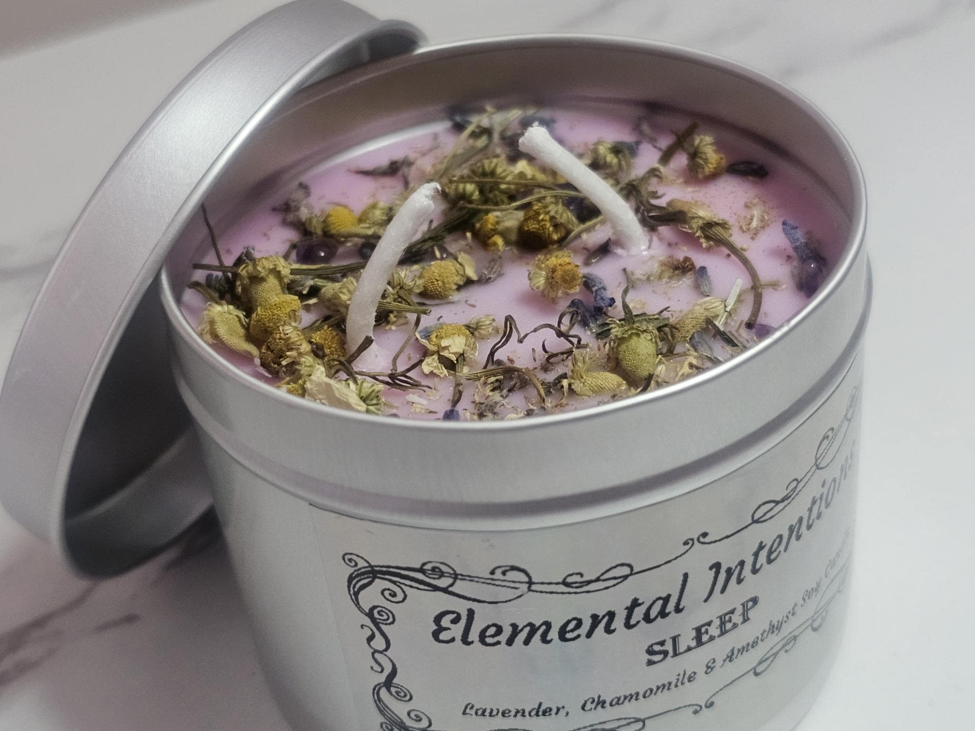 A slightly above shot of a silver container candle with a lid behind it and a light purple candle, adorned with chamomile pieces and small crystal chips. The front lable is silver and holographic with an oriental looking frame around the words &#39;Elemental Intentions.&#39; &#39;Sleep&#39; and the at the bottom of the label are the words &#39;Lavender, Chamomile and Amethyst&#39; soy wax candle.