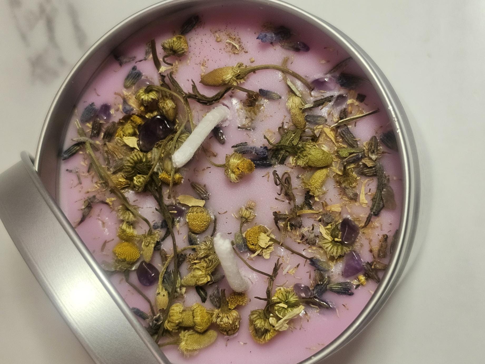 An above shot of a handmade soy wax candle in light purple topped with decorative chamomile pieces and small purple amethyst crystal chips, on a white table.