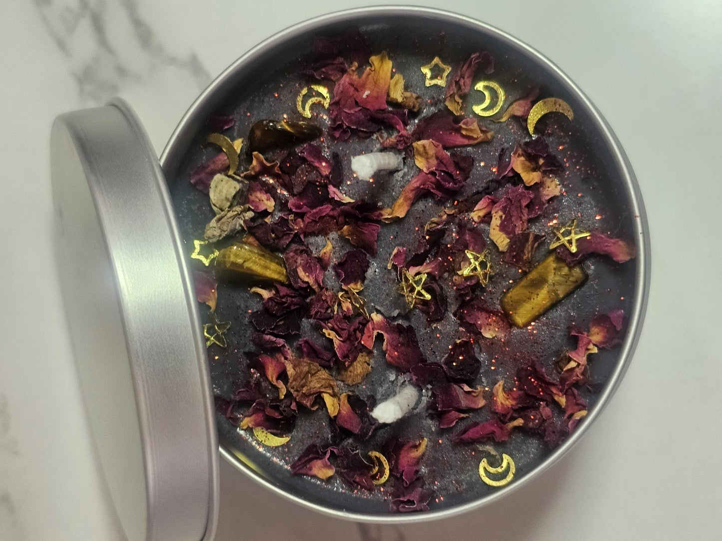 An above shot of a silver container with a black candle topped with rose petals, two white cotton wicks and gold star and moon decorative pieces. Advertised as a decorative &#39;Hecate&#39; tribute scented candle.
