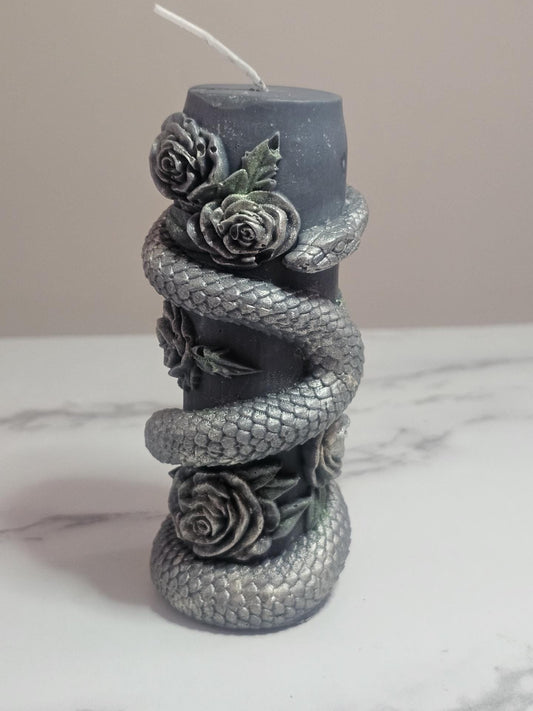 A black pillar candle with a 3D snake winding around the candle, brushed in rose gold candle paint with roses and a leaf deign in between the snake. Painted in rose gold and green with a white cotton wick, on a white marble surface.