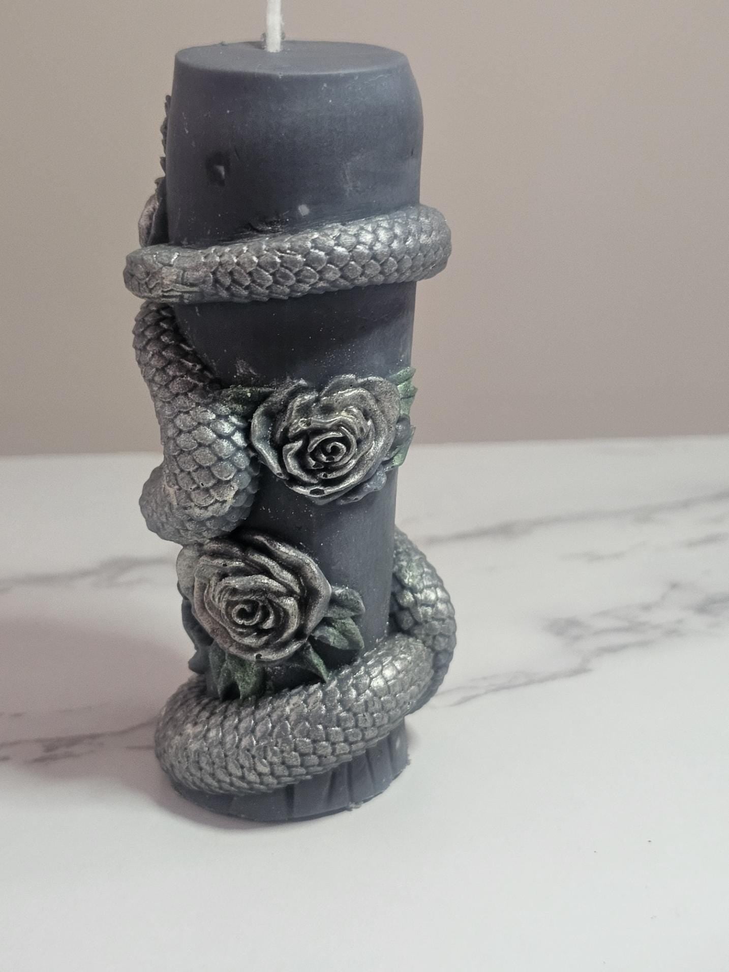 A black pillar altar candle with a 3D snake winding around the candle, brushed in rose gold candle paint with roses and a leaf deign in between the snake. Painted in rose gold and green with a white cotton wick, on a white marble surface.