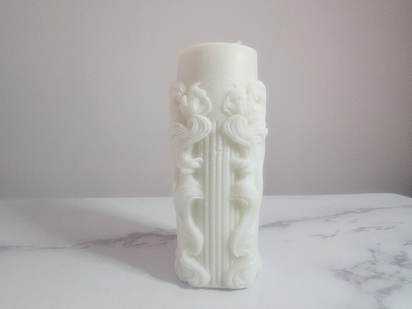 A white pillar altar candle with framed columns all the way around and white cotton wick on a white marble shelf with a cream background.