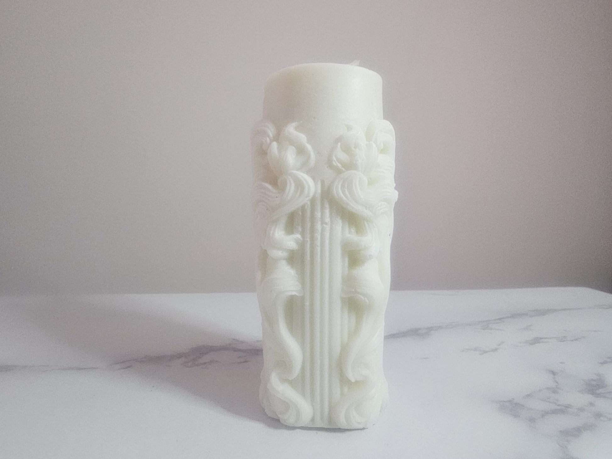 A white pillar altar candle with framed columns all the way around and white cotton wick on a white marble shelf with a cream background.
