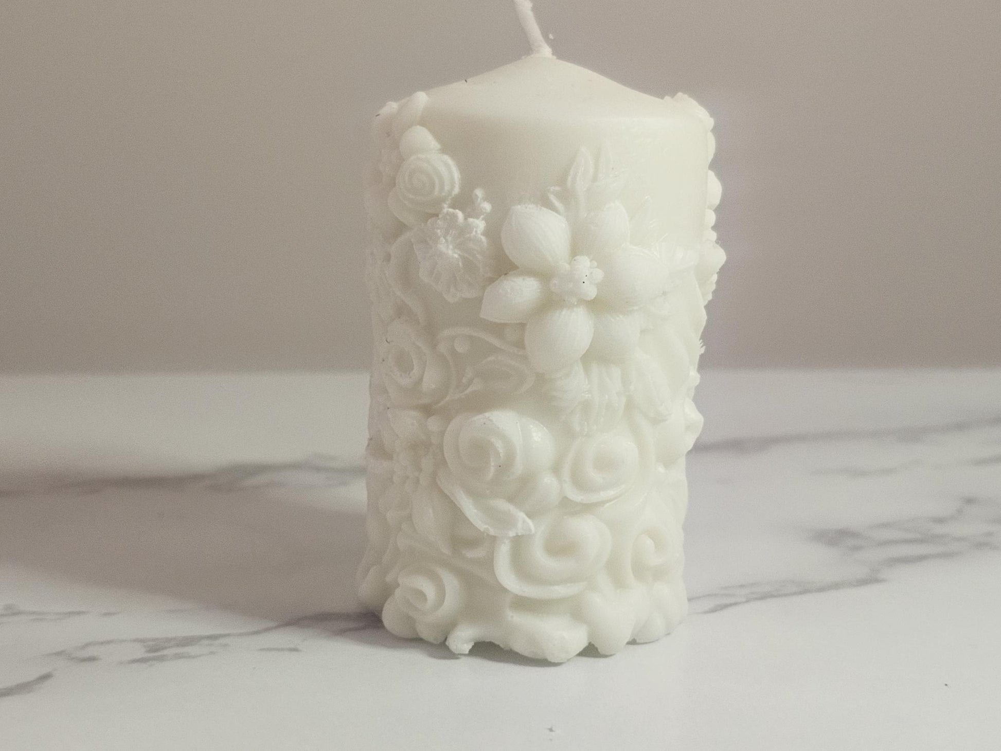 A white soy wax pillar altar candle adorned in floral 3D etchings all the way around with flowers, roses and leaf design and a cotton wick.