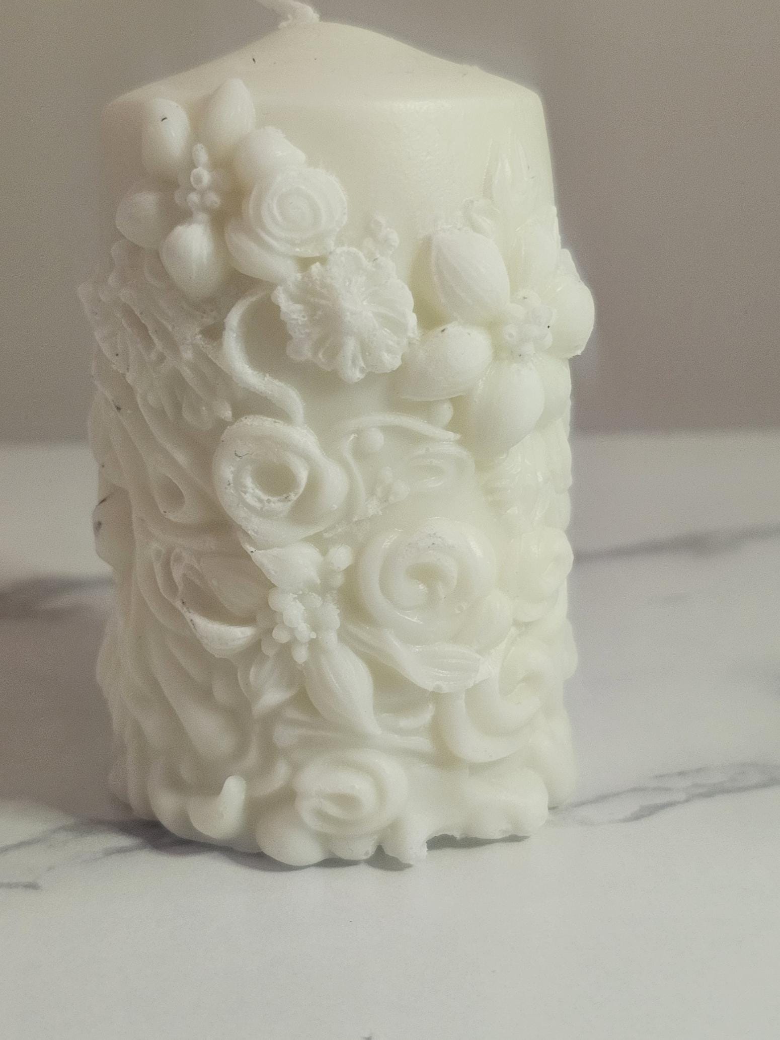 A white soy wax pillar altar candle adorned in floral 3D etchings all the way around with flowers, roses and leaf design and a cotton wick.