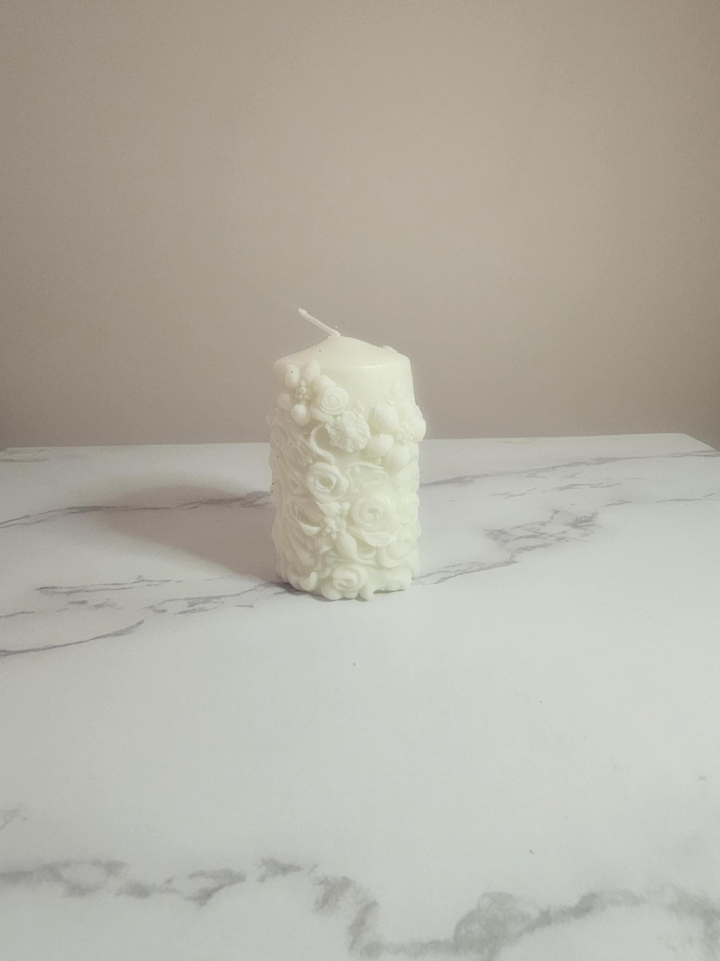 A white soy wax pillar candle adorned in floral 3D etchings all the way around with flowers, roses and leaf design and a cotton wick placed on a marble table  with a cream background.