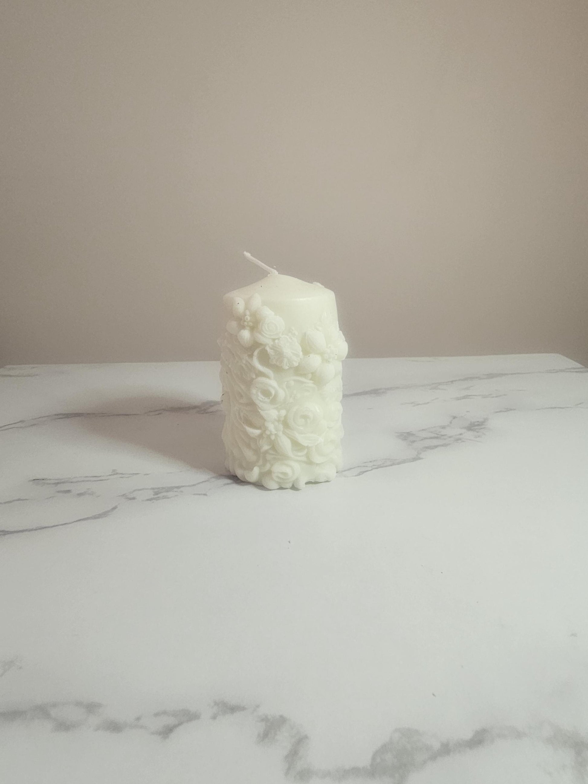 A white soy wax pillar candle adorned in floral 3D etchings all the way around with flowers, roses and leaf design and a cotton wick placed on a marble table  with a cream background.