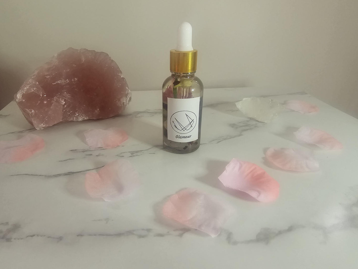 A clear dropper bottle with a gold lid, filled with oil and pink flowers and herbs. There is a white label on the front with a sigil and it is named &#39;glamour&#39;. It is on a white background surrounded by pink rose petals and a rose quartz crystal.