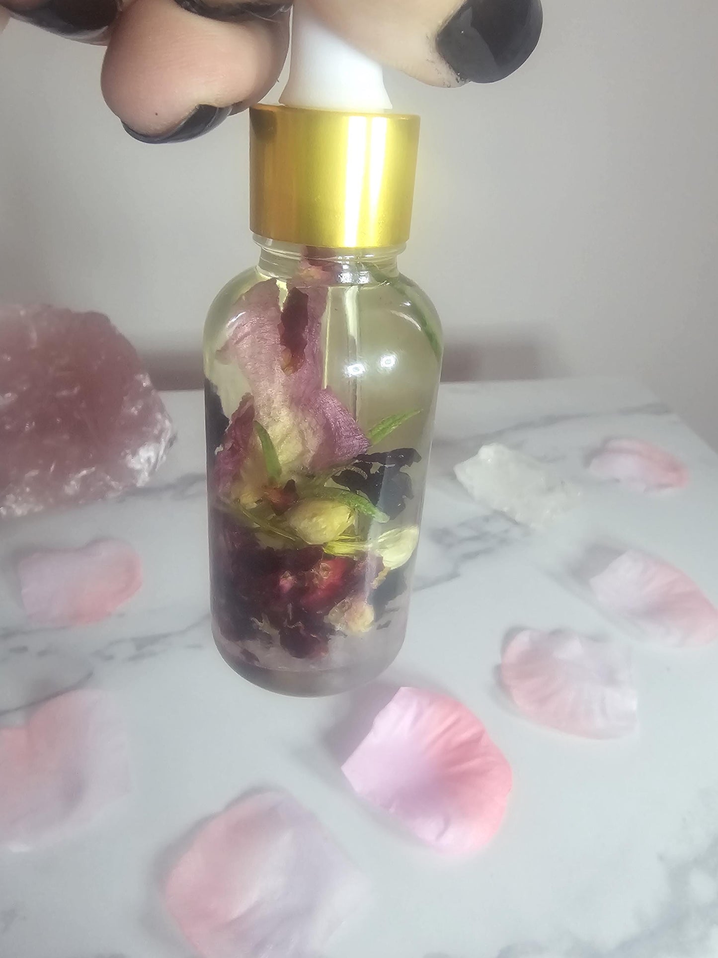 A hand with black nails holding a dropper bottle with a gold top. The bottle is filled with petals, herbs and oil. The bottle is surrounded by pink petals and a large pink crystal.