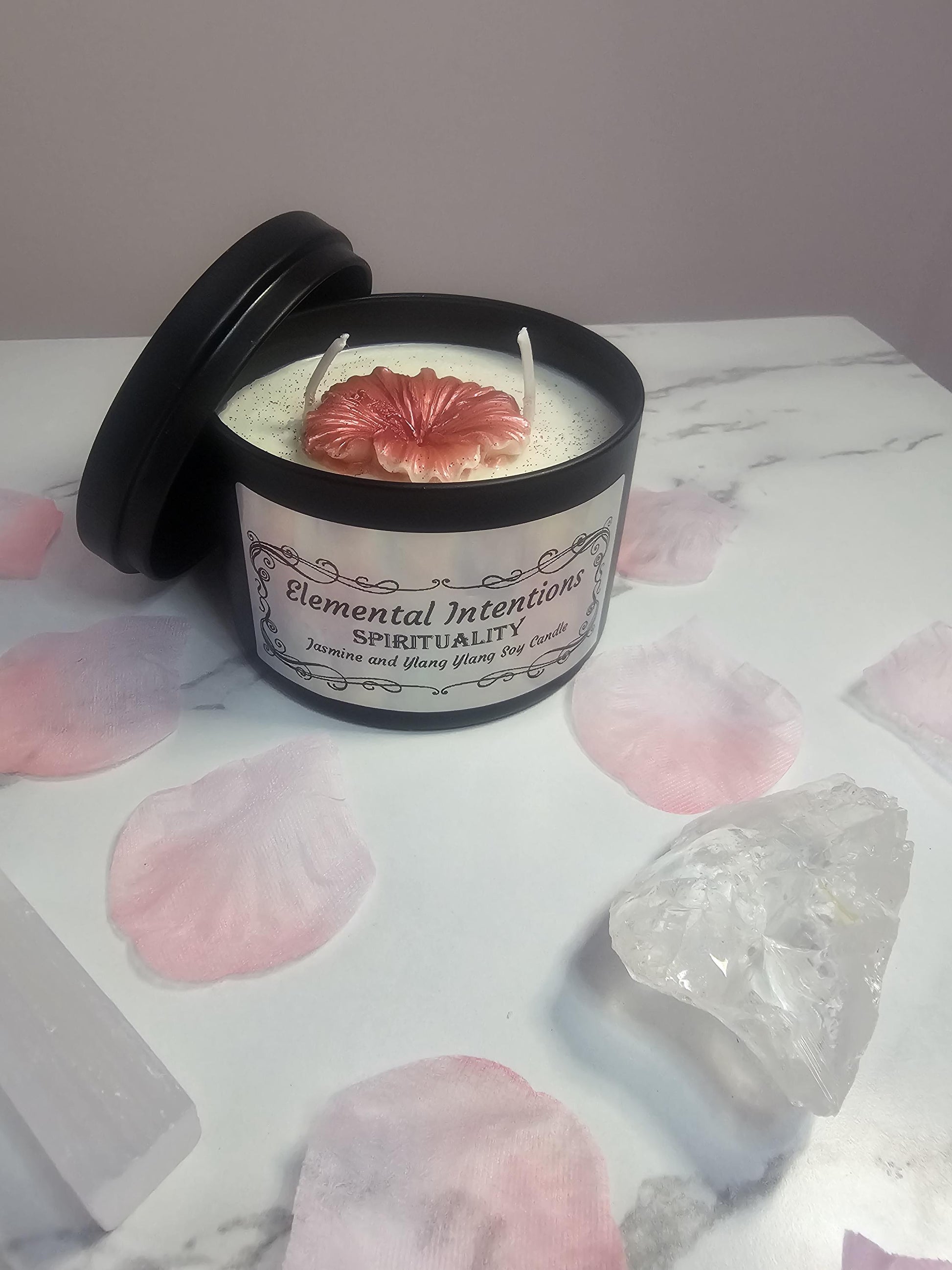 A black candle container filled with white soy wax and a pink peony candle on the top. The front has a label in holographic silver which says &#39;elemental intentions&#39;, &#39;spirituality&#39; and &#39;jasmine and ylang ylang soy candle&#39;. It&#39;s surrounded by pink petals and a clear quartz crystal.