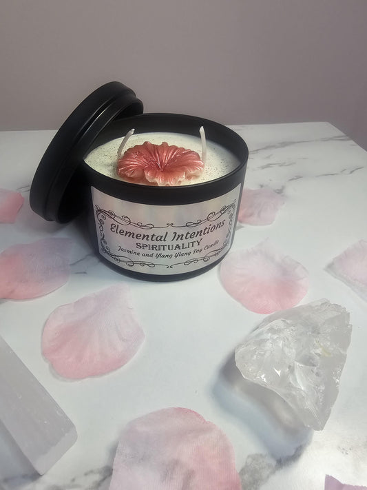 A black candle container filled with white soy wax and a pink peony candle on the top. The front has a label in holographic silver which says &#39;elemental intentions&#39;, &#39;spirituality&#39; and &#39;jasmine and ylang ylang soy candle&#39;. It&#39;s surrounded by pink petals and a clear quartz crystal.