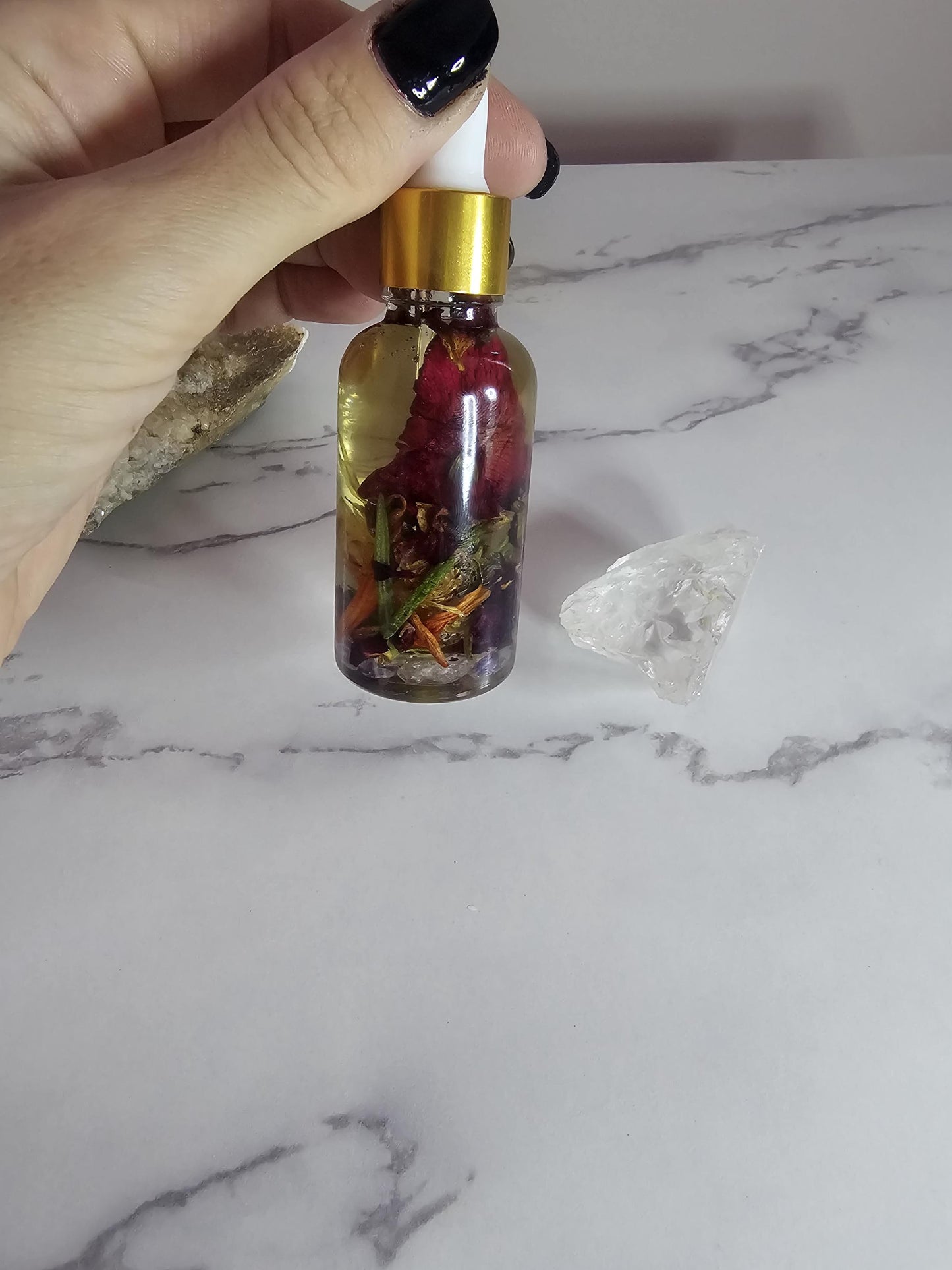 A hand with black glass nails holding a clear glass dropper bottle with a gold top. The inside is filled with clear oil and colourful red and orange flower petals, as well as herbs and small crystals.