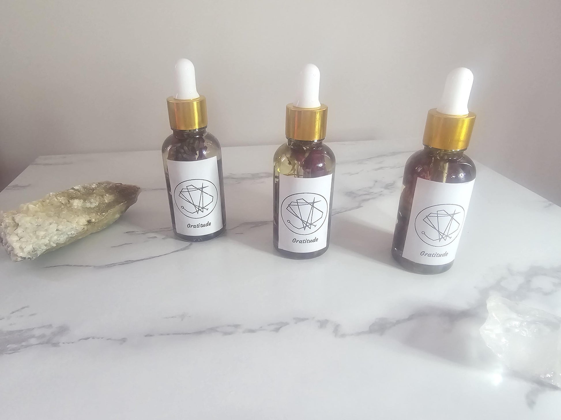 A row of 3 clear glass dropper bottles with god tops. The front labels have a sigil and also say &#39;gratitude&#39;. They are surrounded by crystals on a white background.